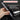 A hand holds a compact gray pen-style tool with a metal tip against a dark background, with marketing text reading 'COMPACT PEN DESIGN' and additional copy about slim aluminum alloy housing for repairing electronics. A small white-and-gray accessory case is shown at lower right.
