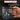 A hand holds a black ceramic mug printed with various hand tools including wrenches, pliers, and screwdrivers in silver and blue. The background is dark and blurred, with promotional text overlays at the top.