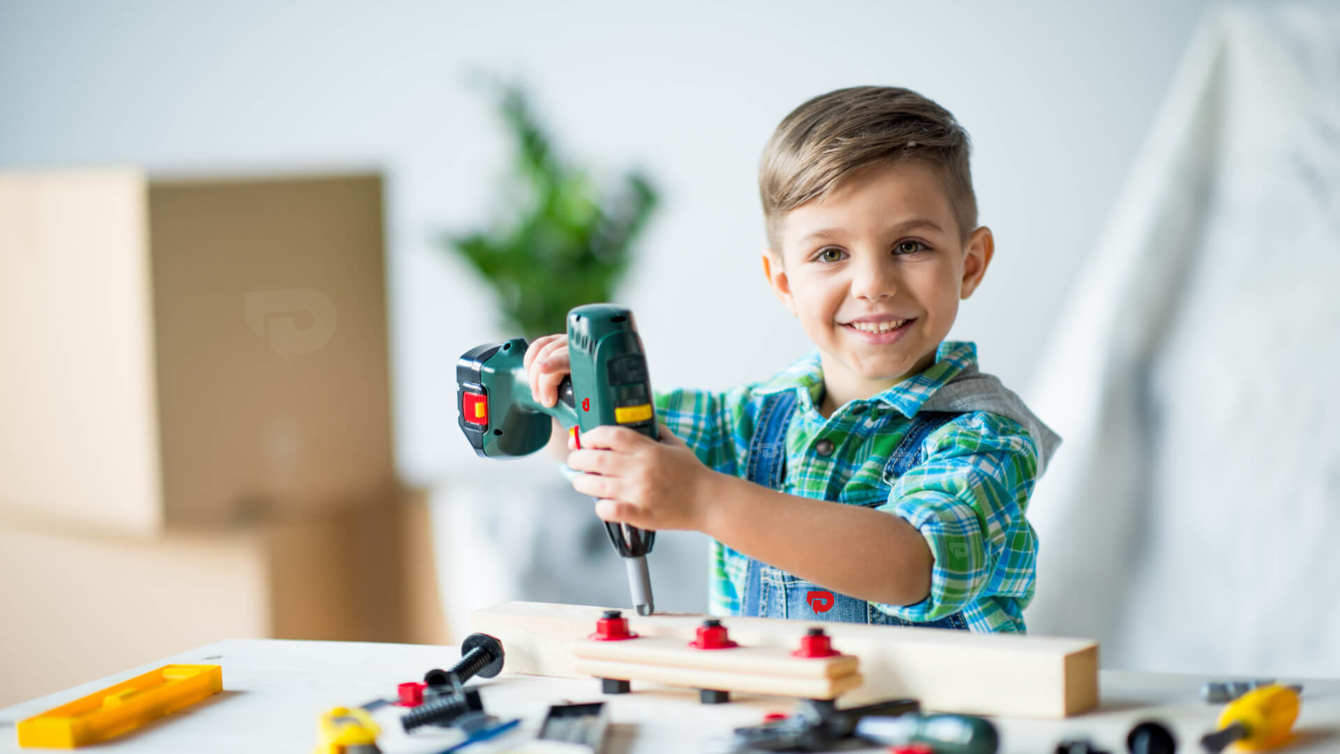 A smiling young boy wearing a green plaid shirt uses a cordless power drill on a wooden project, surrounded by various hand tools and fasteners. The background features moving boxes and a houseplant, suggesting a home improvement or DIY learning activity