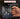 A hand holds a black ceramic mug printed with various hand tools including wrenches, pliers, and screwdrivers in silver and blue. The background is dark and blurred, with promotional text overlays at the top.
